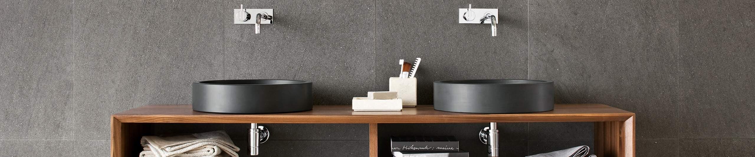 Two black ceramic vessel sinks on wooden vanity against gray tile wall